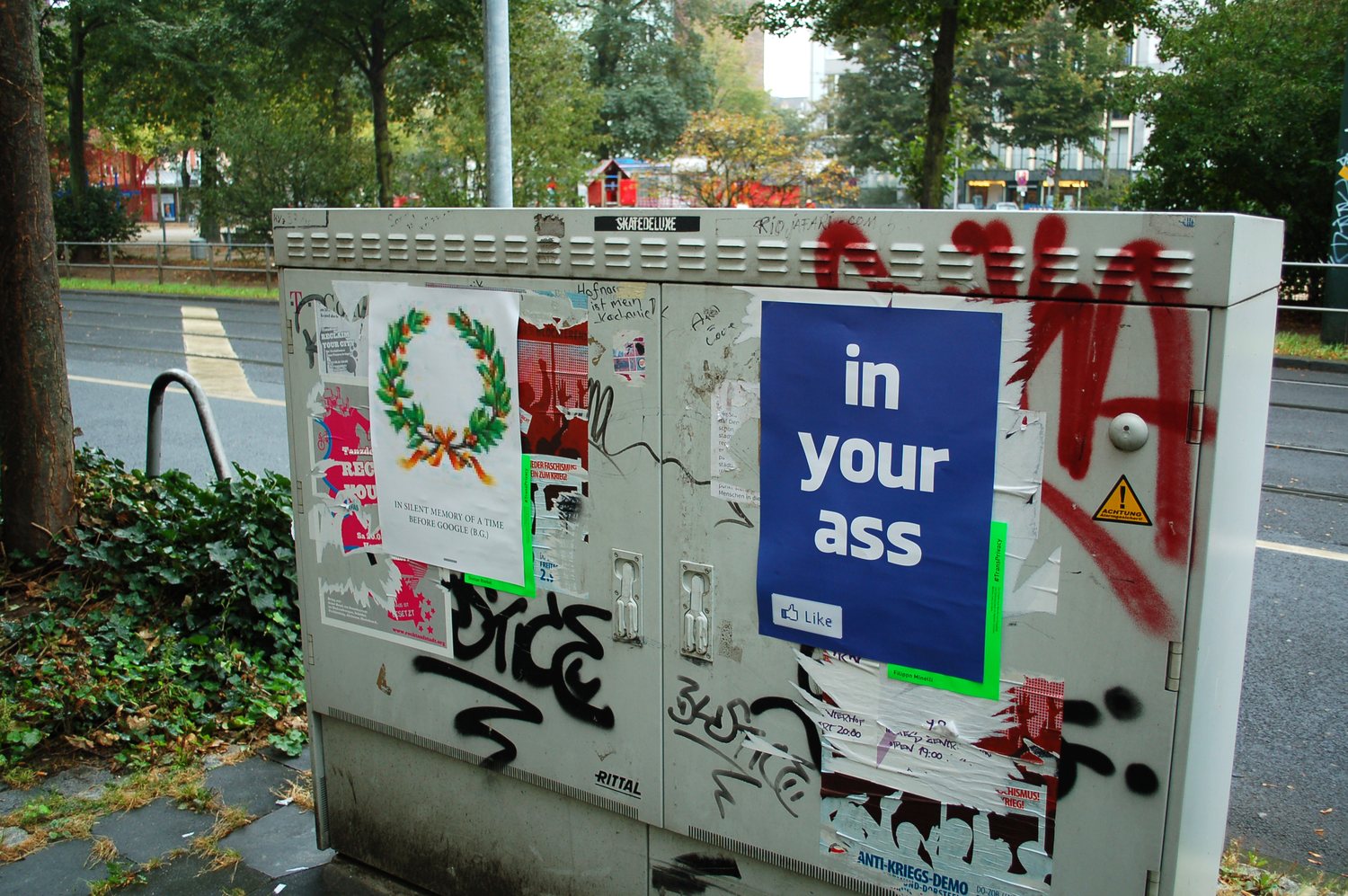 transprivacy — a blue 'in your ass' poster and a laurel-wreath poster on a graffiti-covered utility box